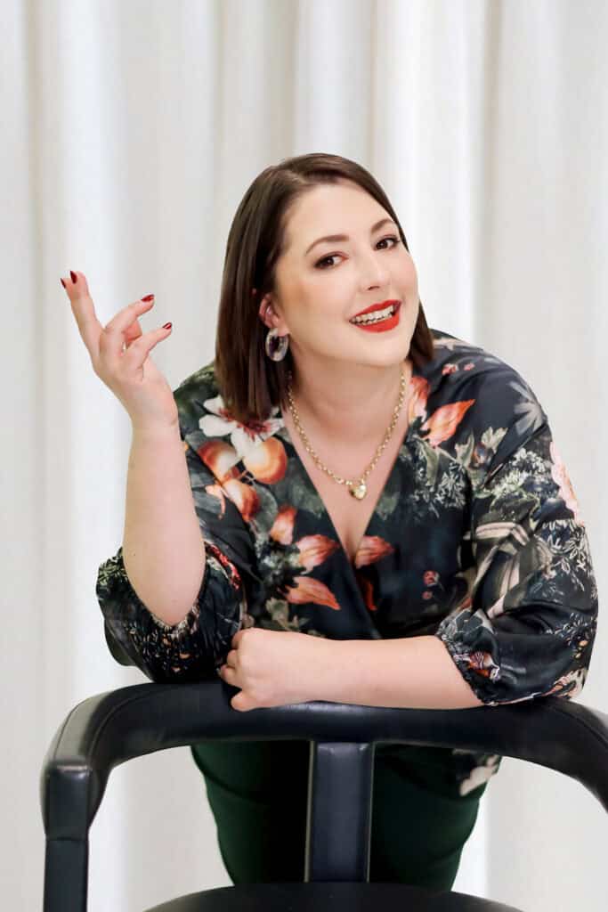 A woman with shoulder-length brown hair and makeup, wearing a floral blouse, sitting on a black chair with a white curtain background, smiling confidently and gesturing with her hand.